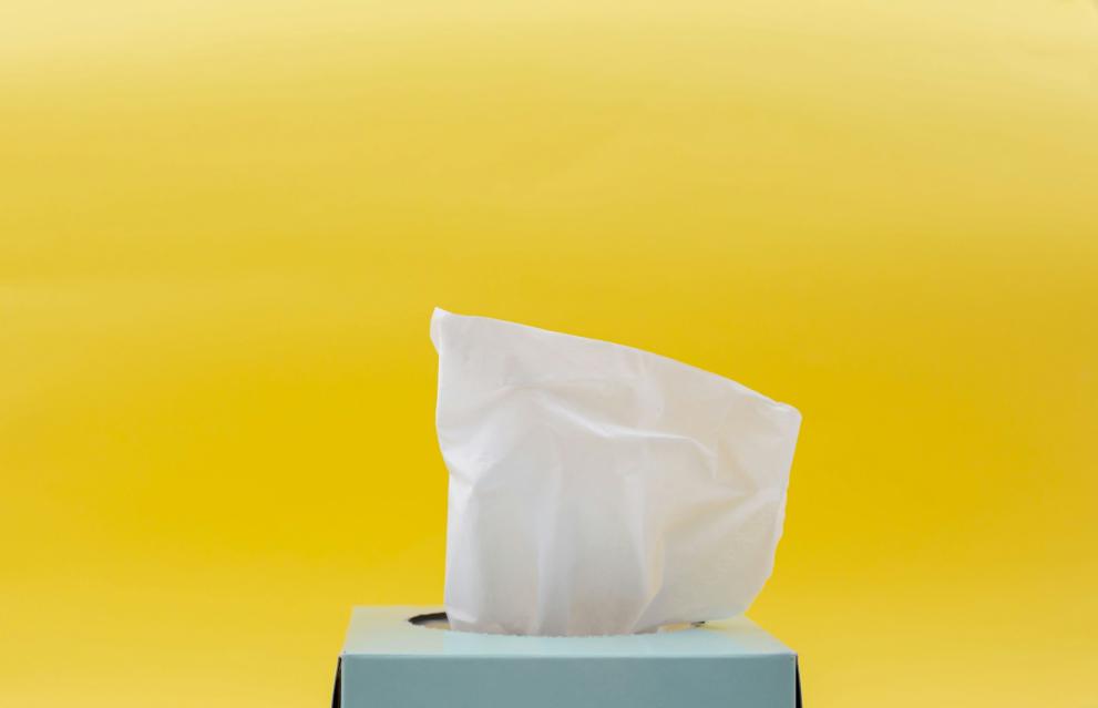 Picture of a light blue tissue box, with a tissue pulled up, on a bright yellow uniform background.